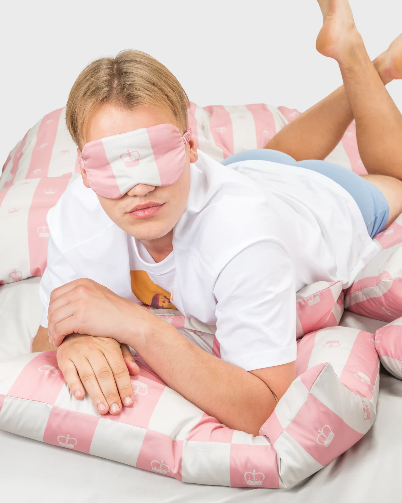Young man with a pink sleep mask lying on pink striped “Your Majesty” bedding