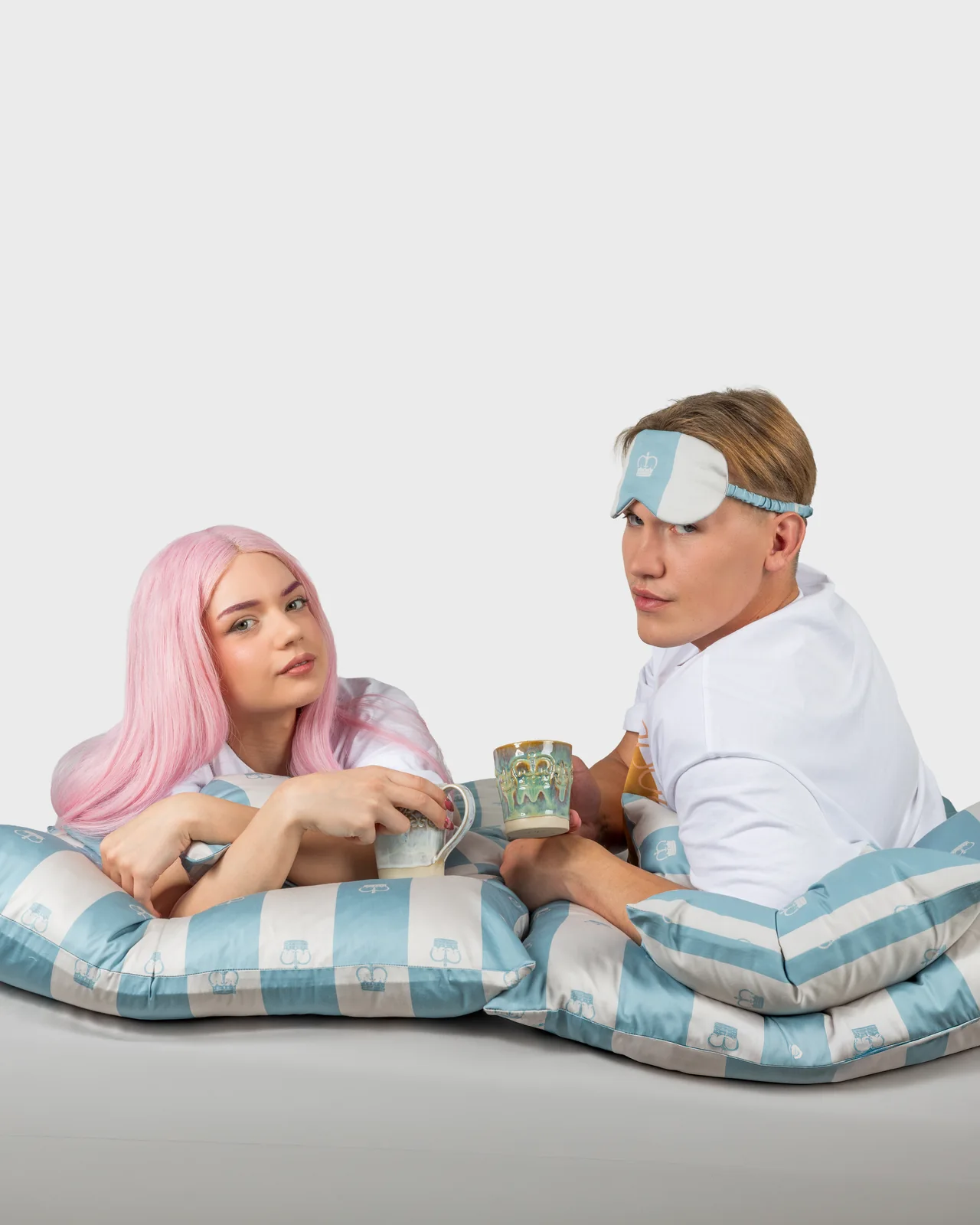 Couple with mugs lying on striped pillows, the young man with a “Your Majesty” sleep mask on his forehead