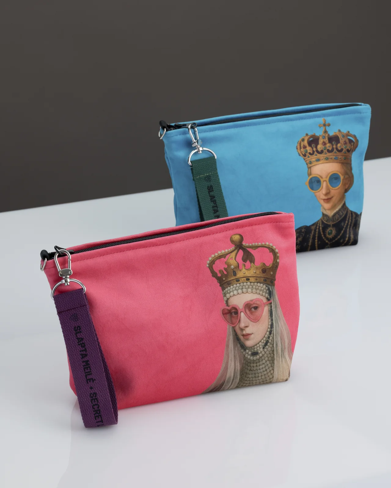 Pink “REBEL QUEEN” cosmetic bag on a white table, a blue cosmetic bag with a queen behind it