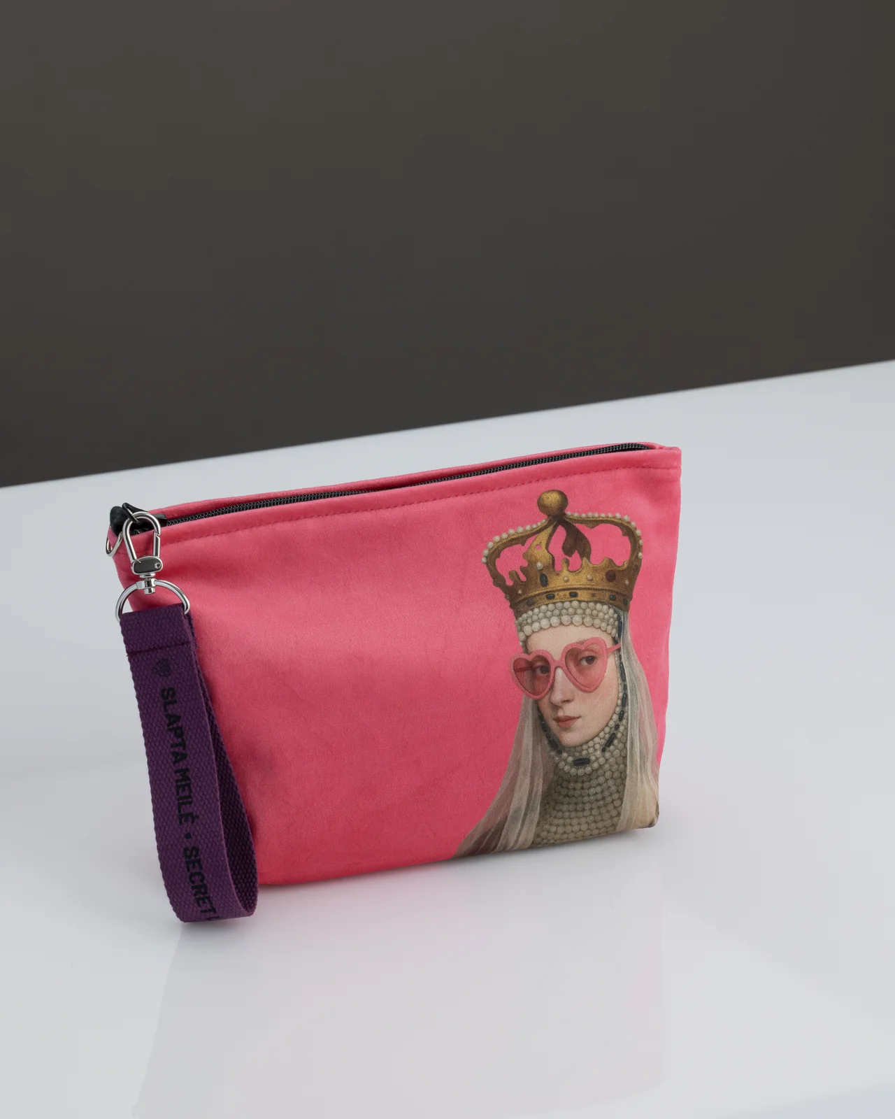Pink “REBEL QUEEN” cosmetic bag with a drawing of a queen and a purple handle on a white table