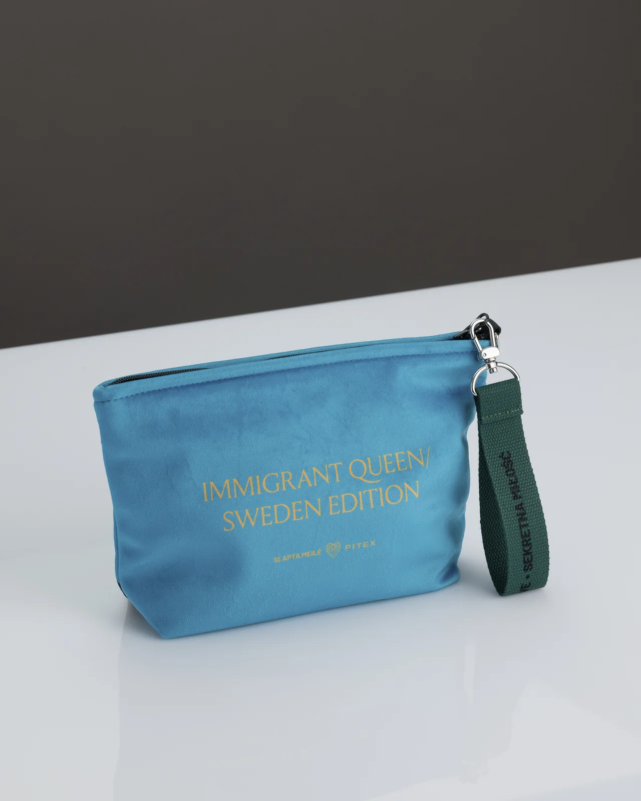 Niebieska kosmetyczka „IMMIGRANT QUEEN: Sweden Edition” na stole, widoczna strona ze złotym napisem