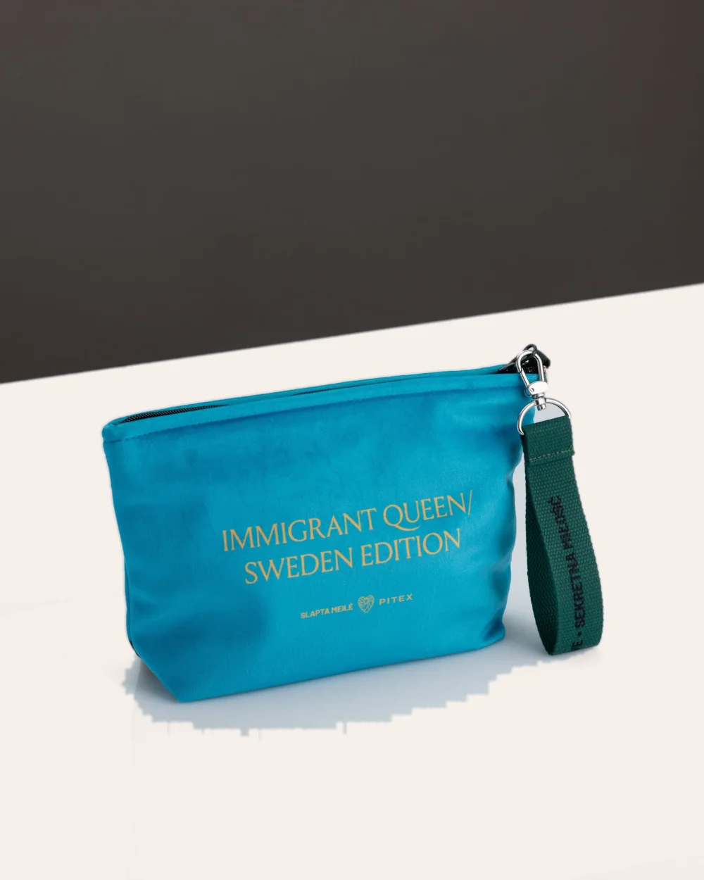 Mėlyna kosmetinė „IMMIGRANT QUEEN: Sweden Edition“ ant stalo, matoma pusė su auksiniu užrašu