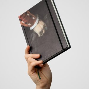 Notes „Slapta meilė | Zygmunt August” z ciemną okładką, uniesiony w dłoni