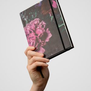 Notes „Slapta meilė | Historie dworskie” z okładką z różowymi kwiatami, uniesiony w dłoni