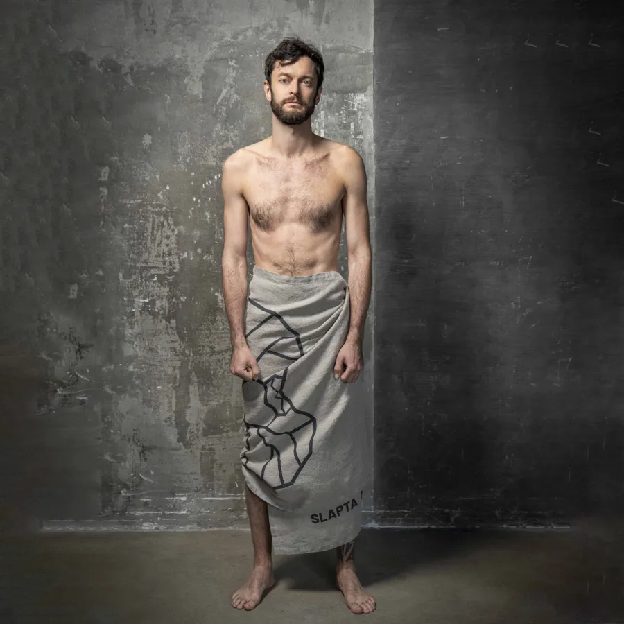 Full-length view of a man with a grey “The King's” linen towel around his hips by a concrete wall
