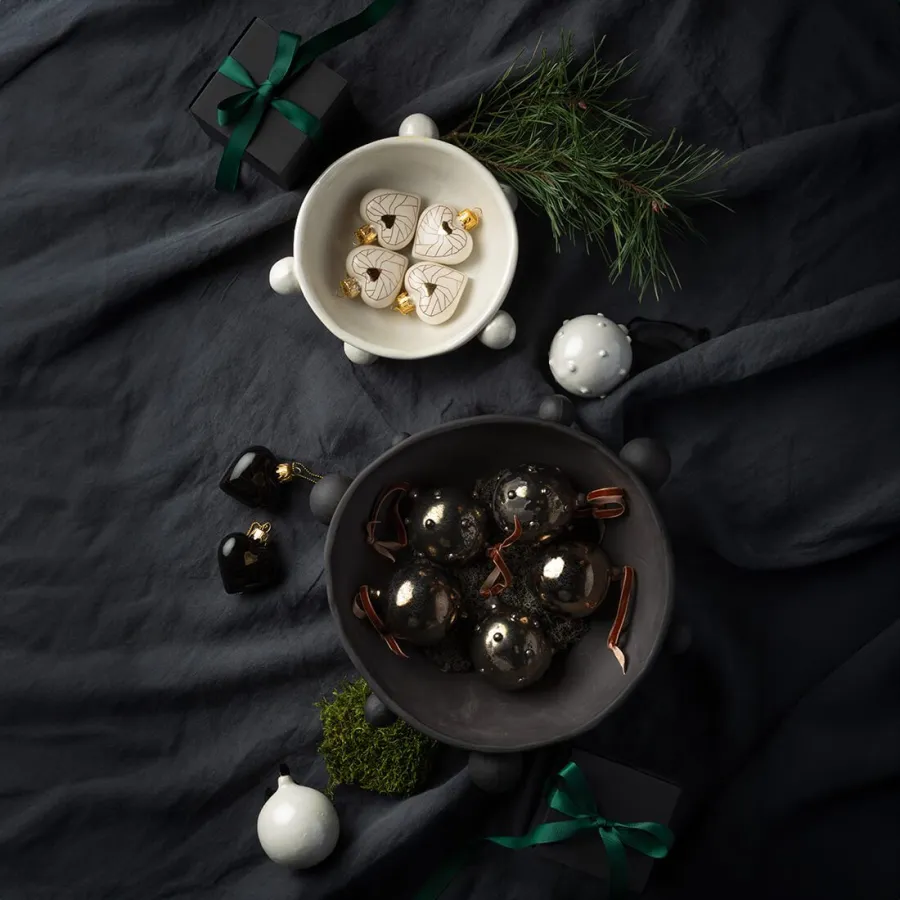 Black and white “Crowned Heart” Christmas ornaments in small bowls on dark fabric with a fir sprig