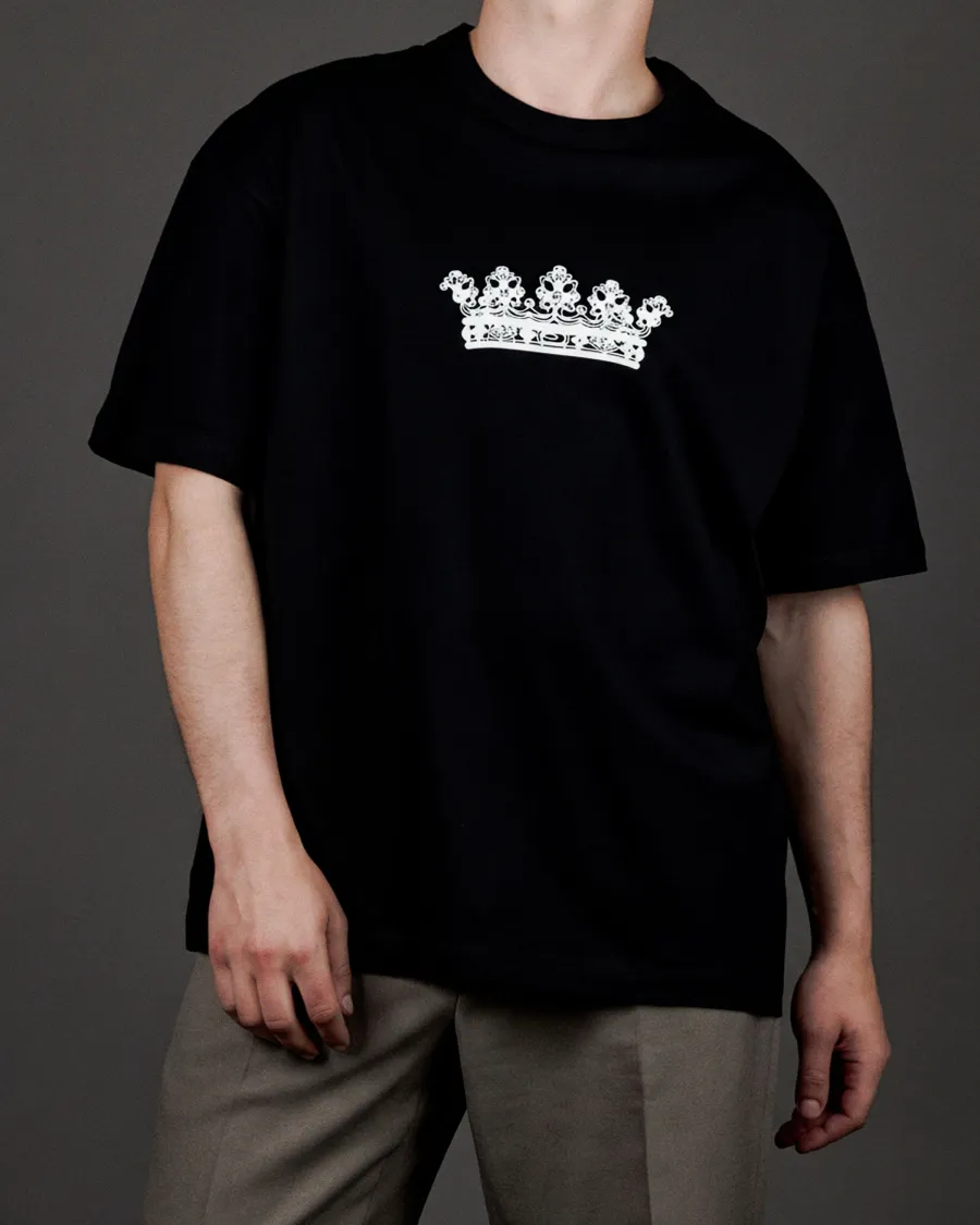 Man wearing a black “KARŪNA” T-shirt with a white crown drawing and grey trousers