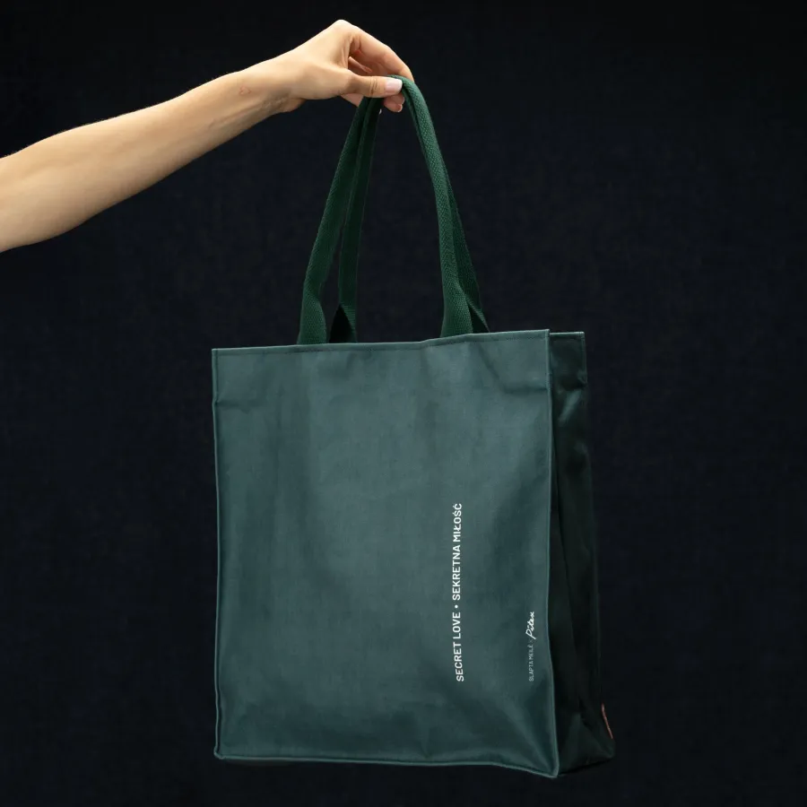 Back of a dark green “Slapta meilė” handbag with lettering, held in a hand on a dark background