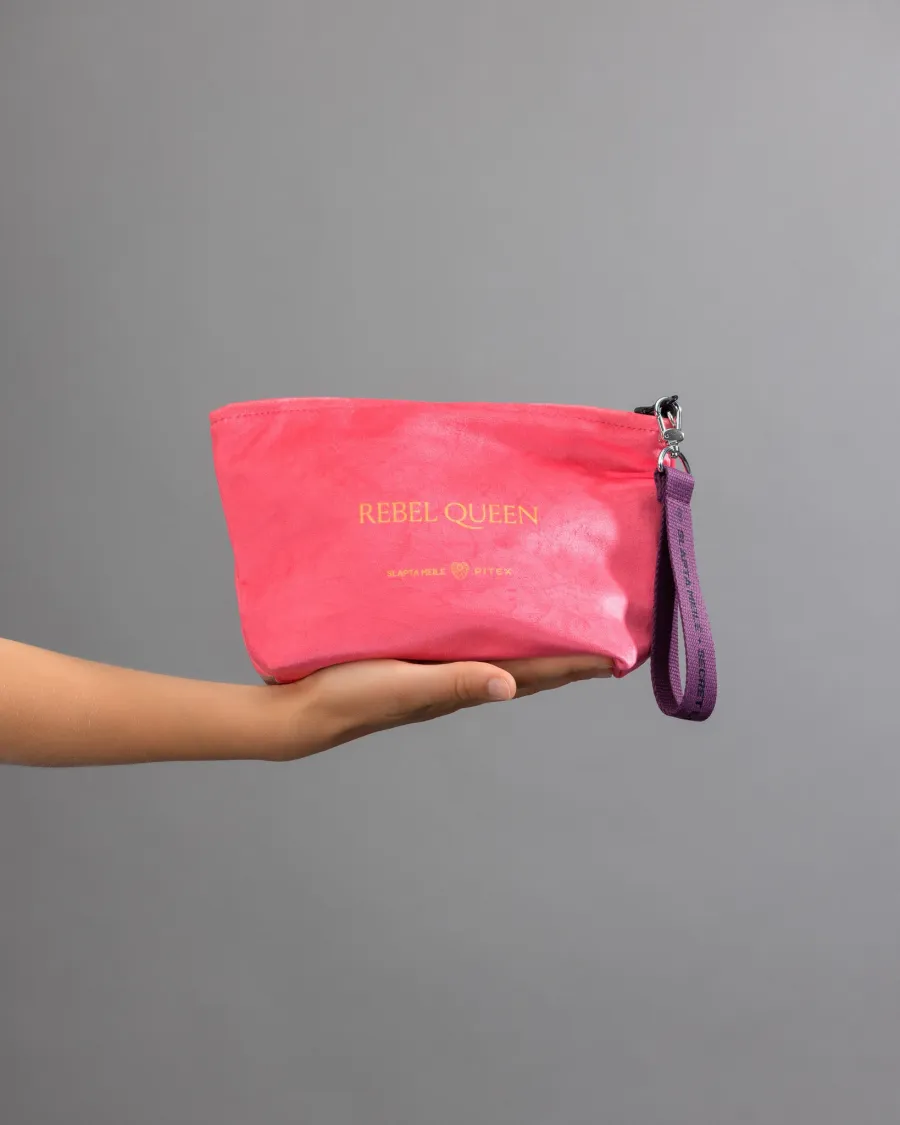 Back of the pink “REBEL QUEEN” cosmetic bag with gold lettering on a palm against a grey background