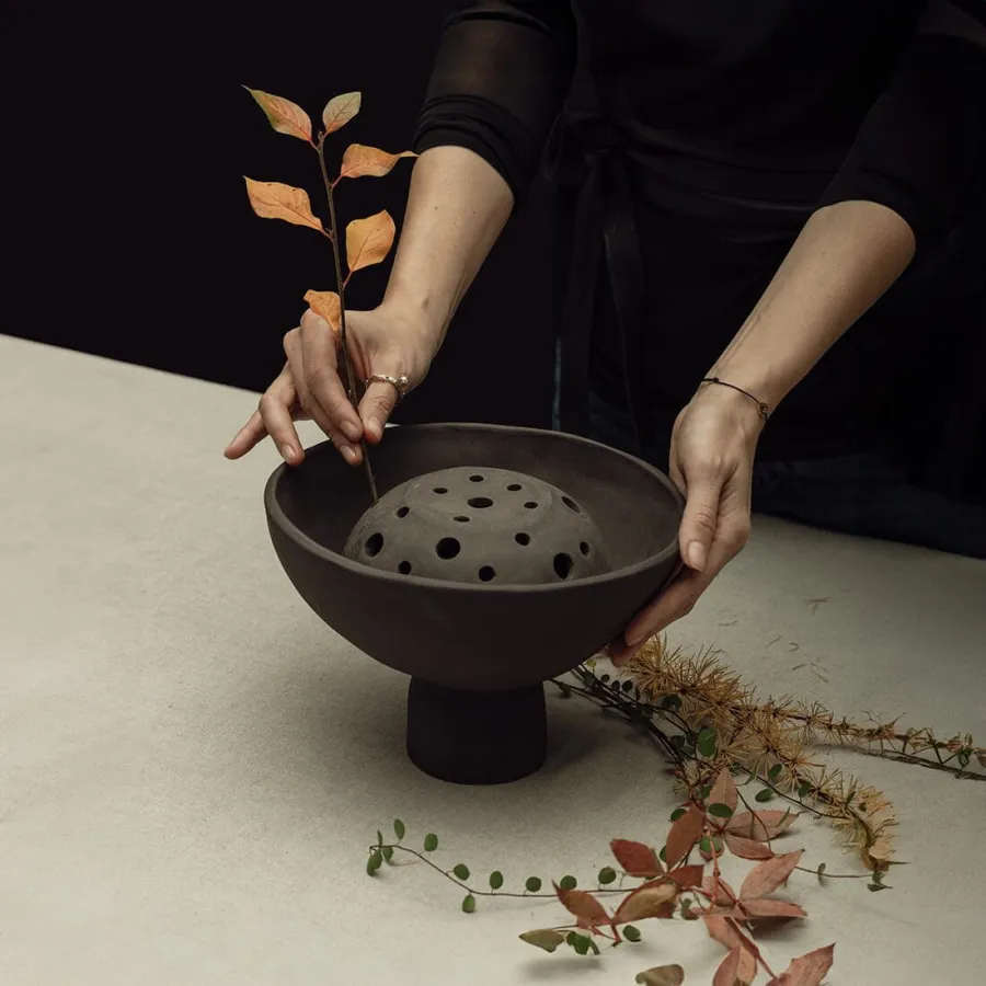 Hands placing autumn twigs into a dark Renaissance-style bouquet vase with holes