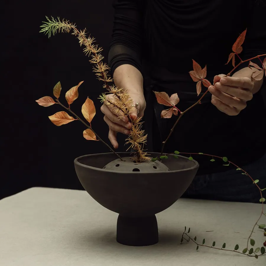 Hands arranging a bouquet of branches in a dark Renaissance-style bouquet vase on a table