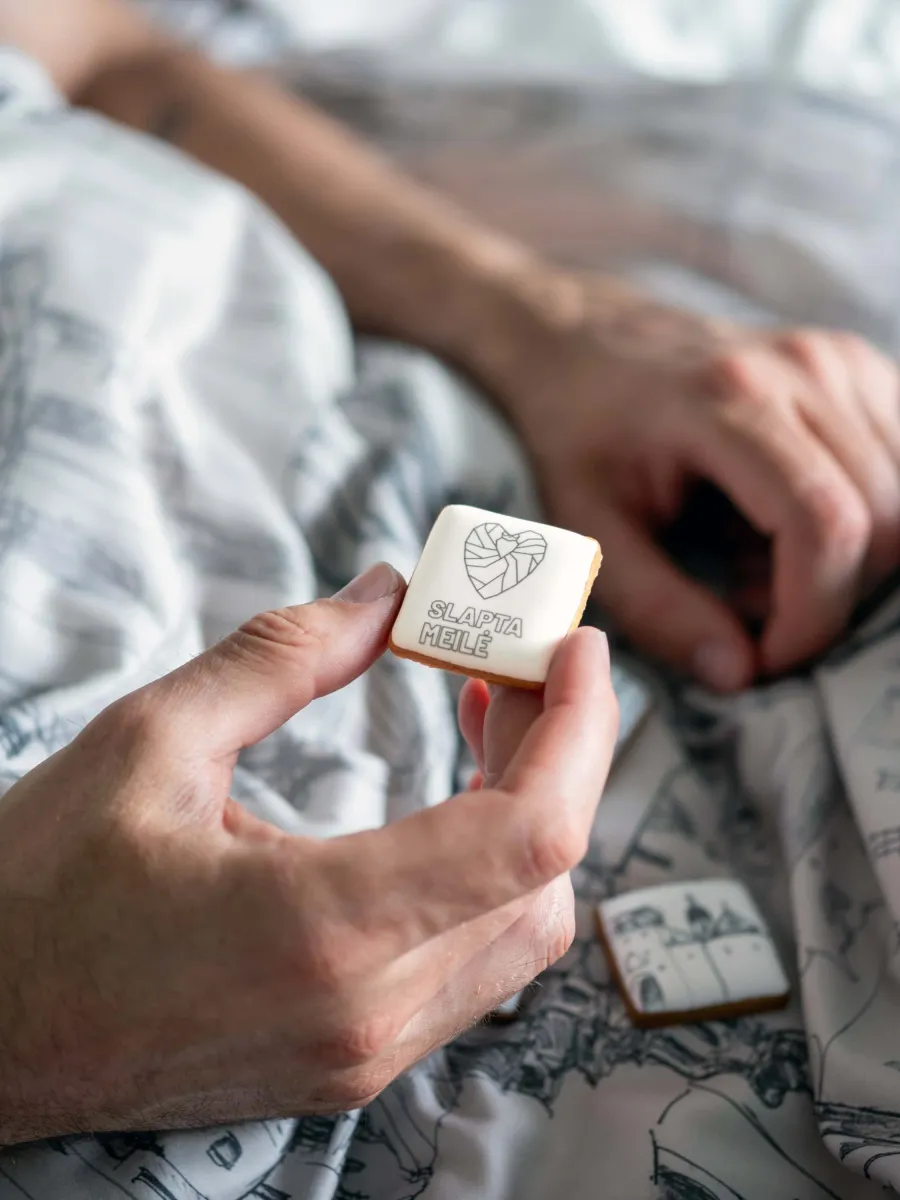 Hand holding a square “Royal Vilnius” gingerbread biscuit with a heart and crown drawing in bed