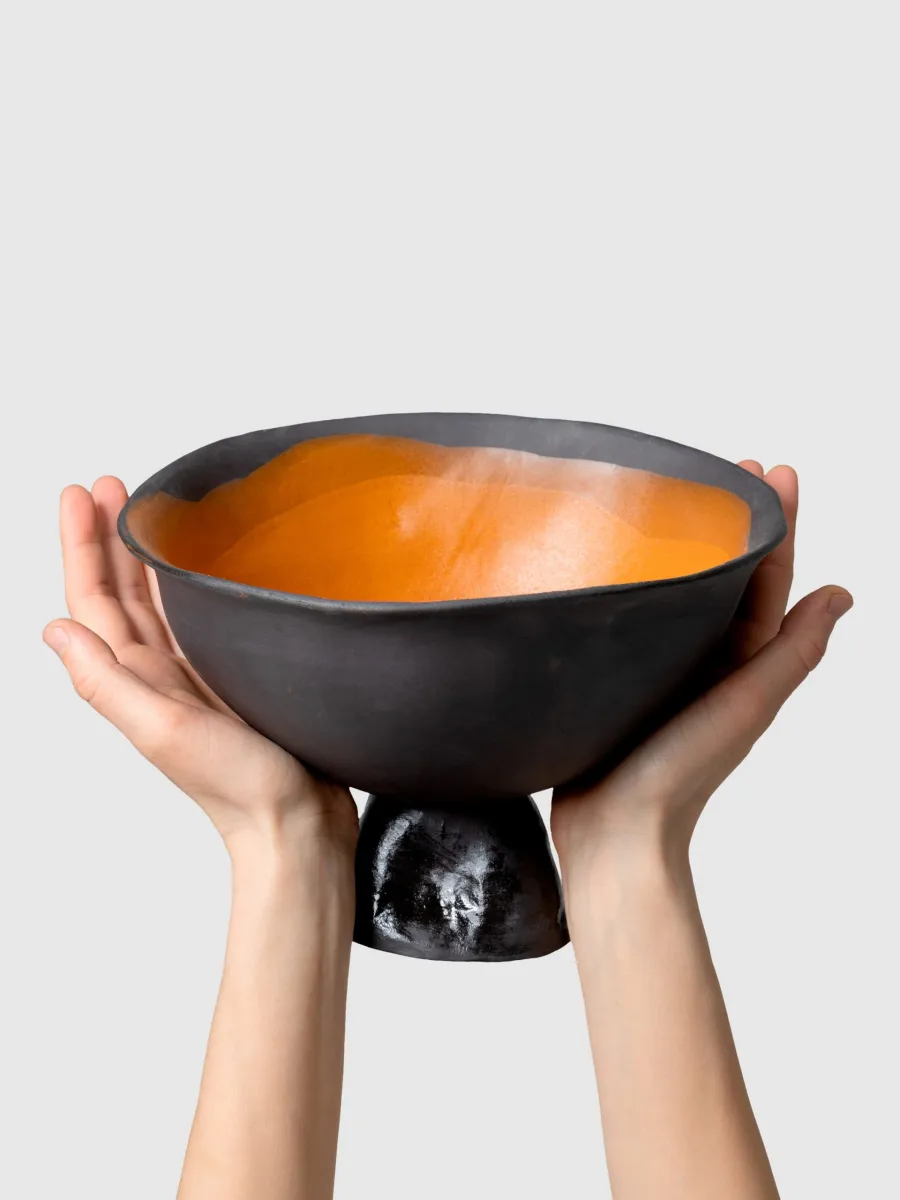 Two hands hold a black Kings' Footed Bowl with an orange inside, front view
