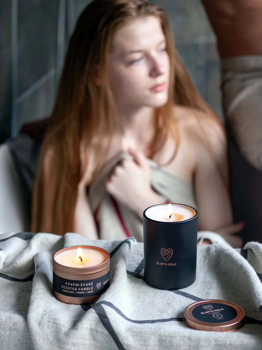 Woman with a blanket next to lit “Slapta meilė” candles and a small candle in a tin