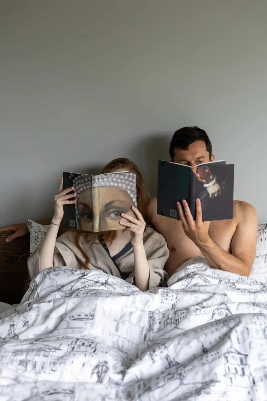 Couple reading books in bed with white satin “Royal Vilnius Love Story” bedding