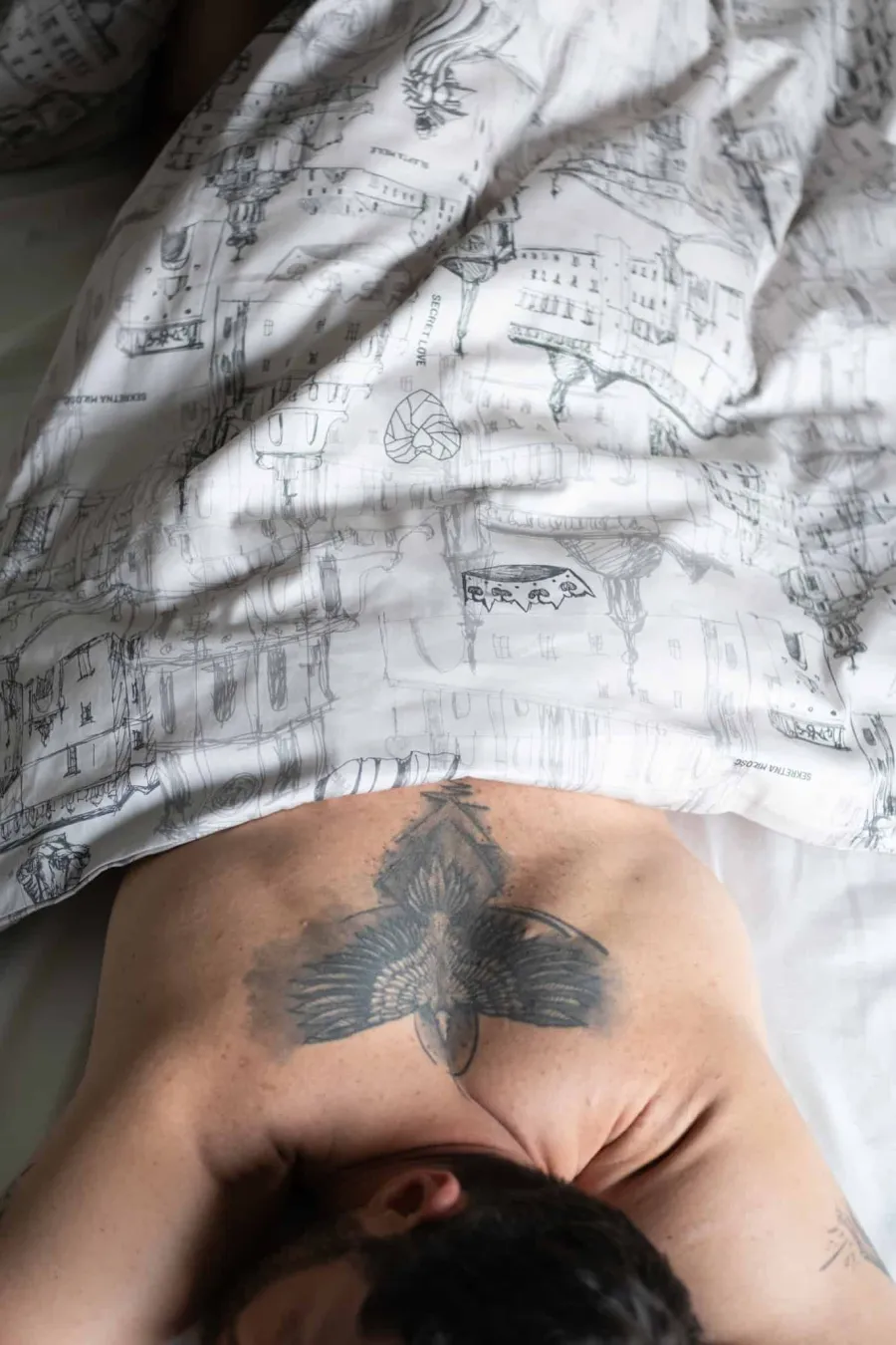 Man with a tattooed back lying under white “Royal Vilnius Love Story” bedding, view from above