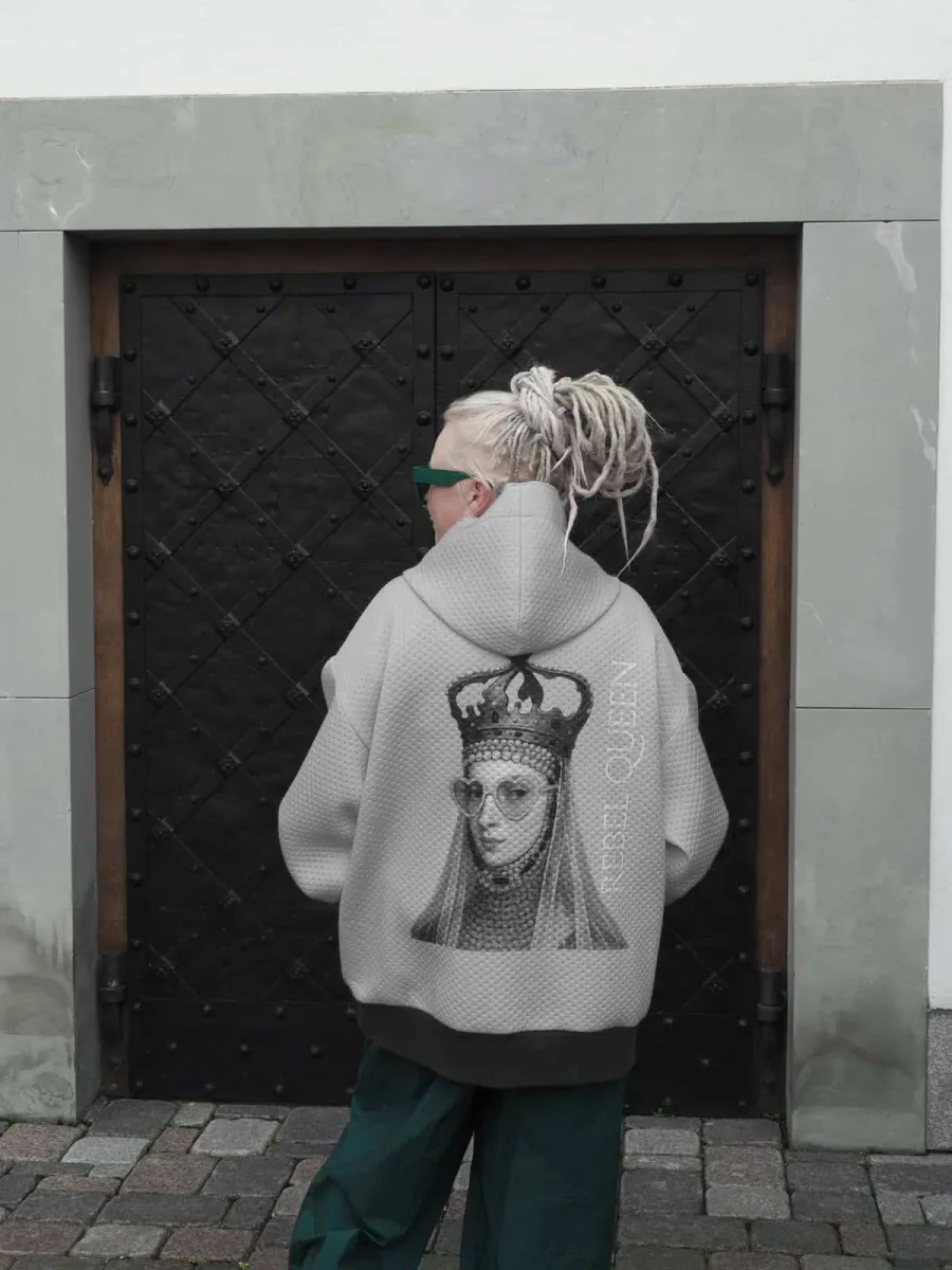 Woman in a grey “Rebel Queen” hoodie with her back to a black door, the queen's image visible