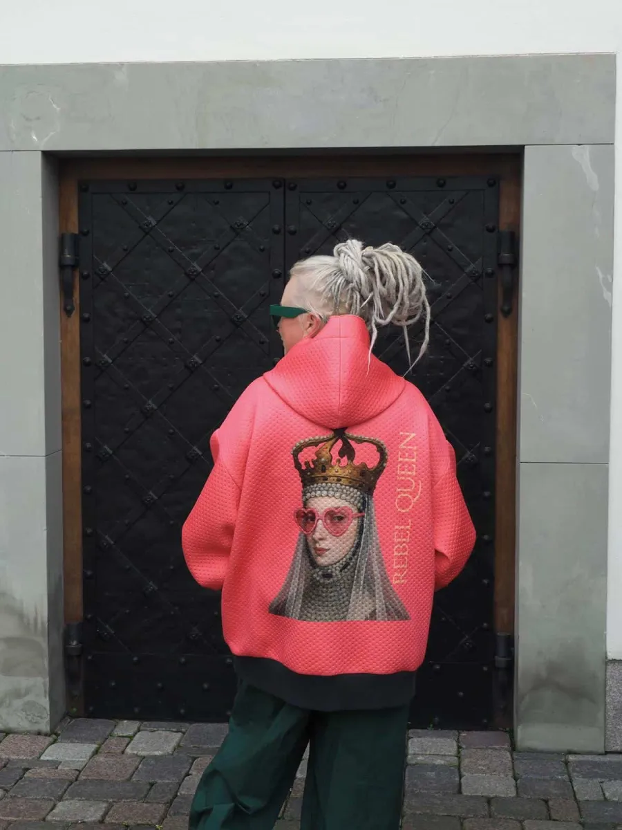 Woman in a coral “Rebel Queen” hoodie with her back to a black door in the street