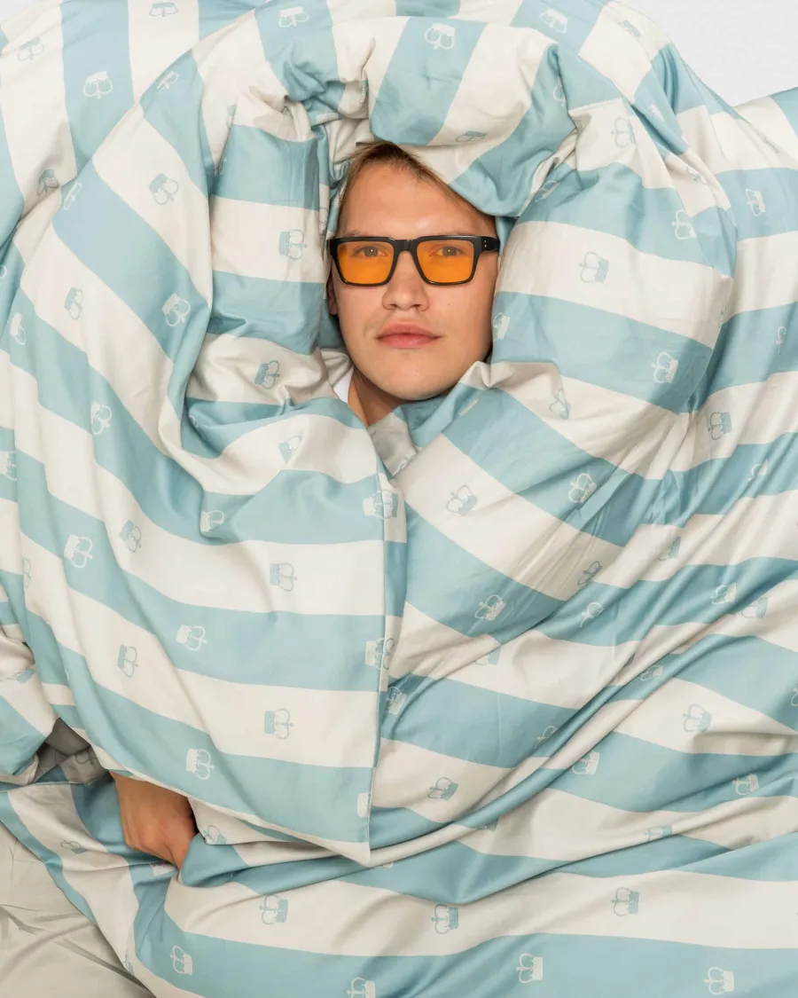 Young man in orange glasses wrapped in a blue and white striped duvet