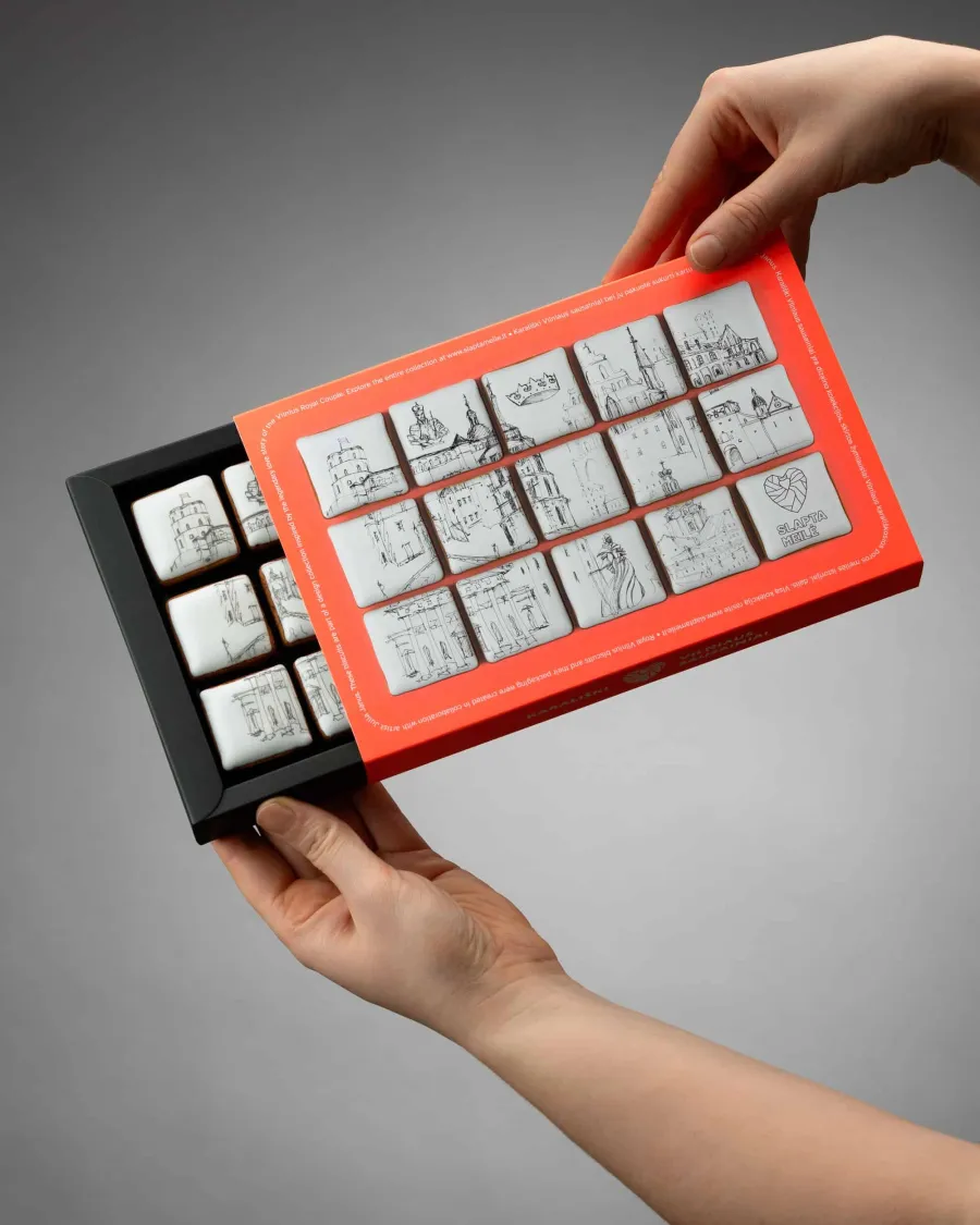 Hands holding an open box of sweets with drawings of Vilnius and a red frame on a grey background