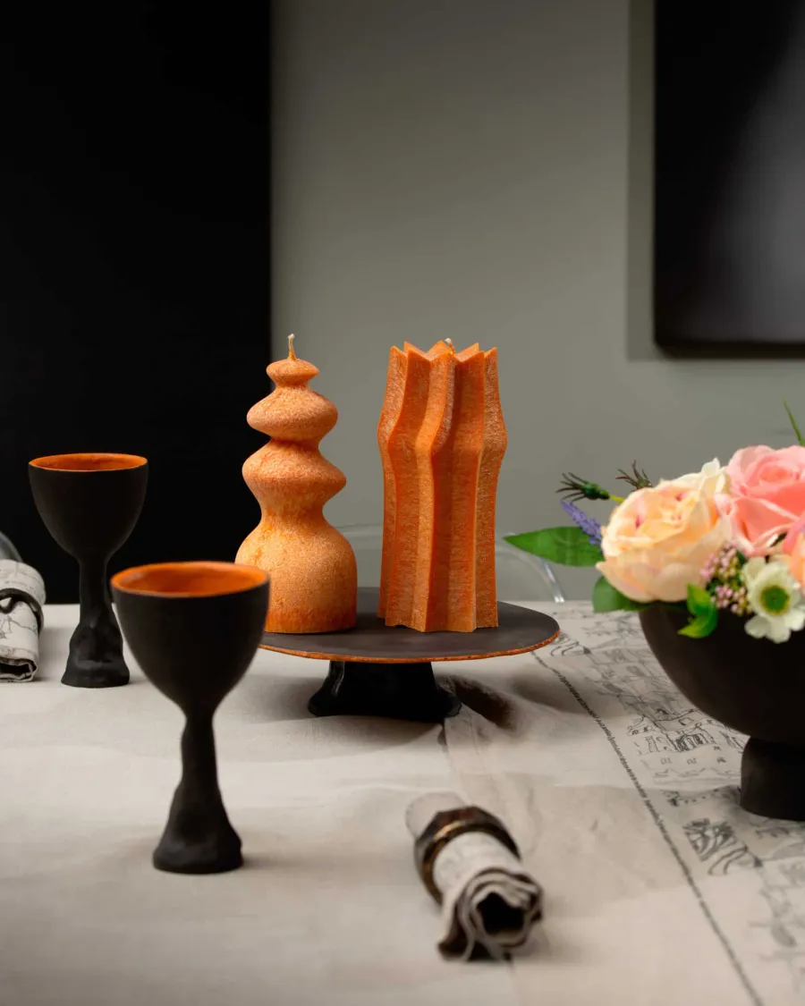 Orange tiered and faceted candles on a black tray next to goblets and a bouquet of roses